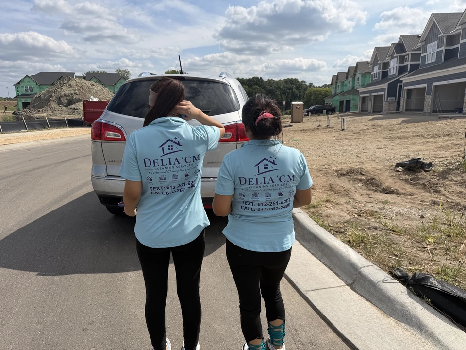Two people seen from behind wearing light blue Delia’cm Cleaning Services shirts beside a vehicle